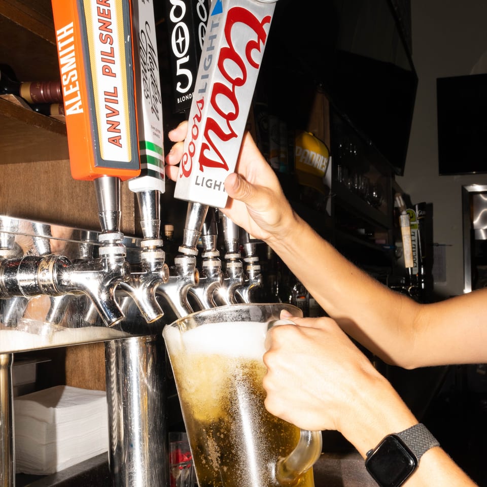 A bartender pours a frothy Coors Light beer from a tap into a large glass pitcher.