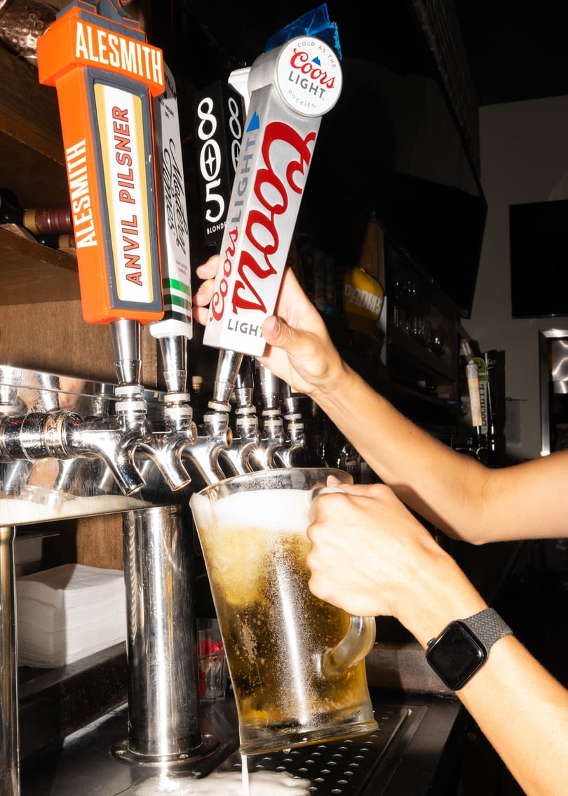 A bartender pours a frothy Coors Light beer from a tap into a large glass pitcher.