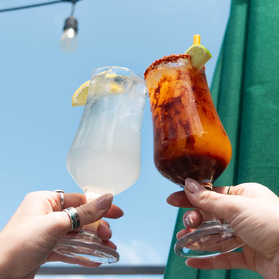 Two hands holding up a light-colored drink and a red-orange drink against a blue sky.