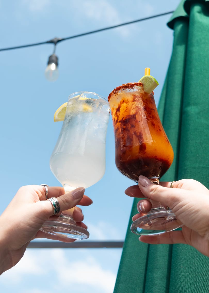 Two hands holding up a light-colored drink and a red-orange drink against a blue sky.