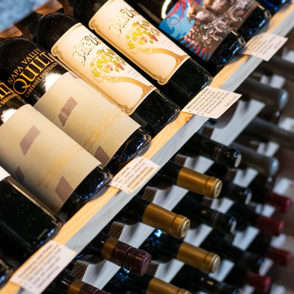 Wine bottles with various labels, including "Napa Valley Quilt" and "Bella Union," displayed on a wooden rack.