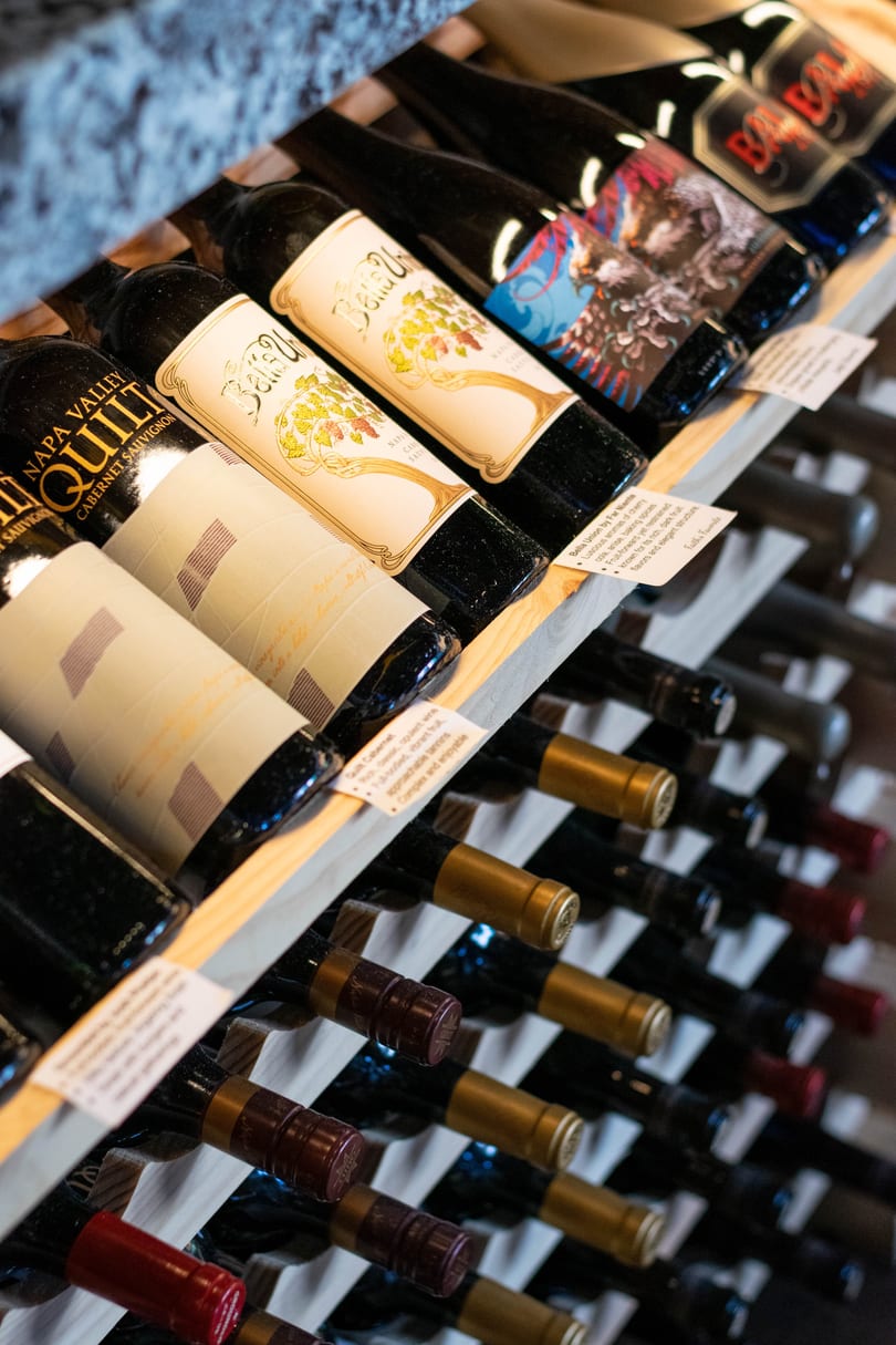 Wine bottles with various labels, including "Napa Valley Quilt" and "Bella Union," displayed on a wooden rack.