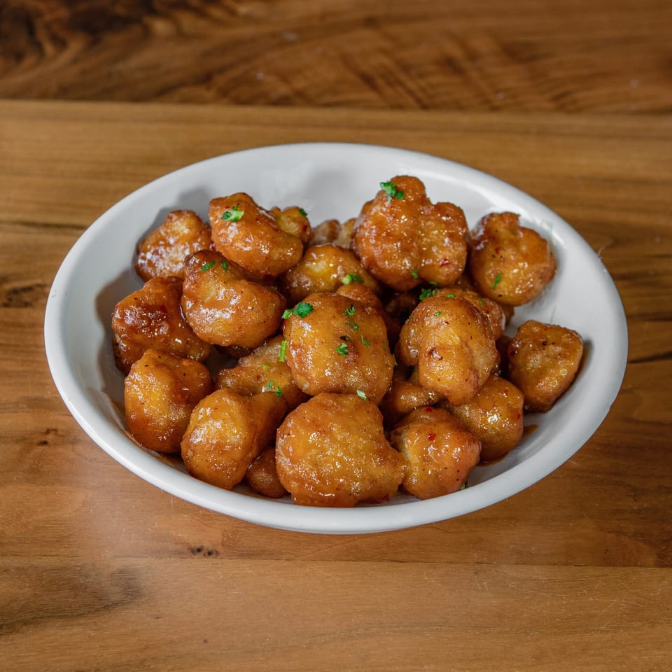 A white bowl filled with golden-brown, glazed popcorn chicken, garnished with green herbs, on a wooden table.