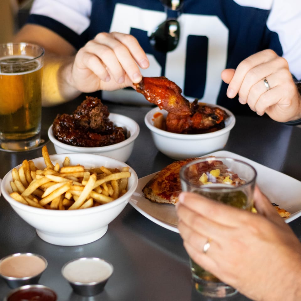 Two people enjoying chicken wings, fries, and beer at a table with various dipping sauces.
