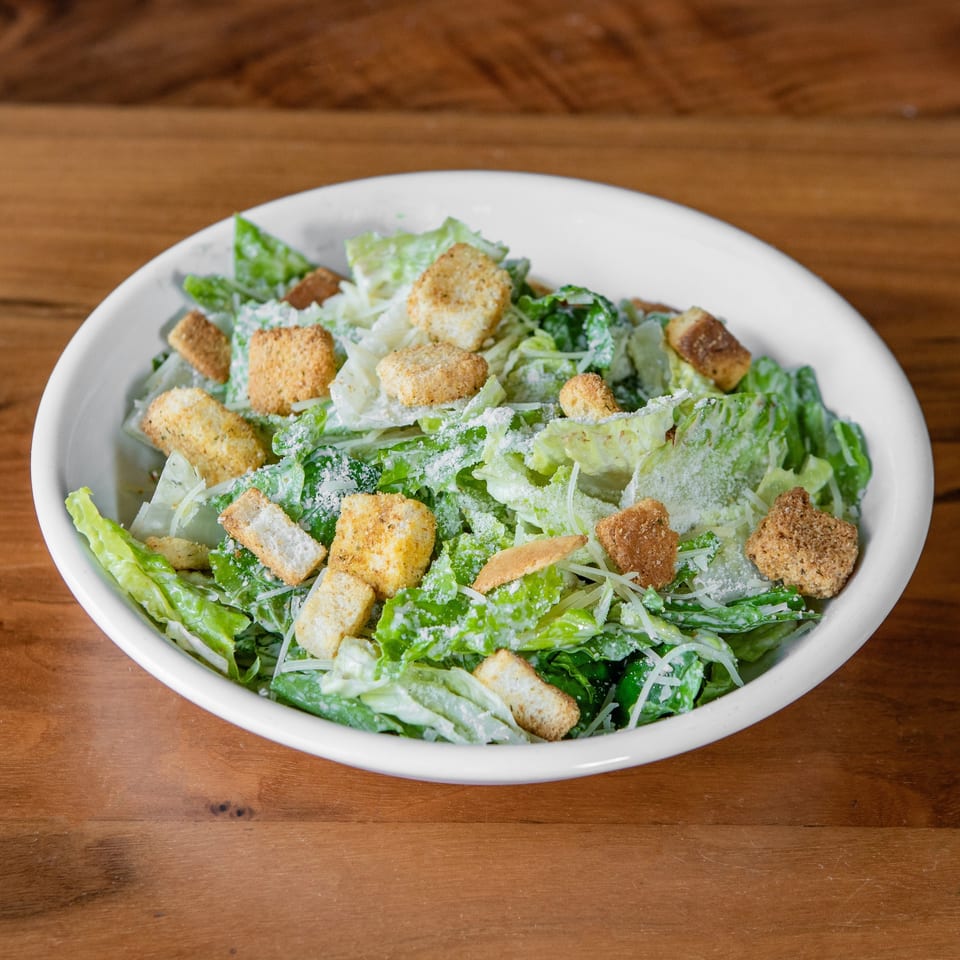 A white bowl filled with a Caesar salad, topped with croutons and grated cheese, on a wooden table.