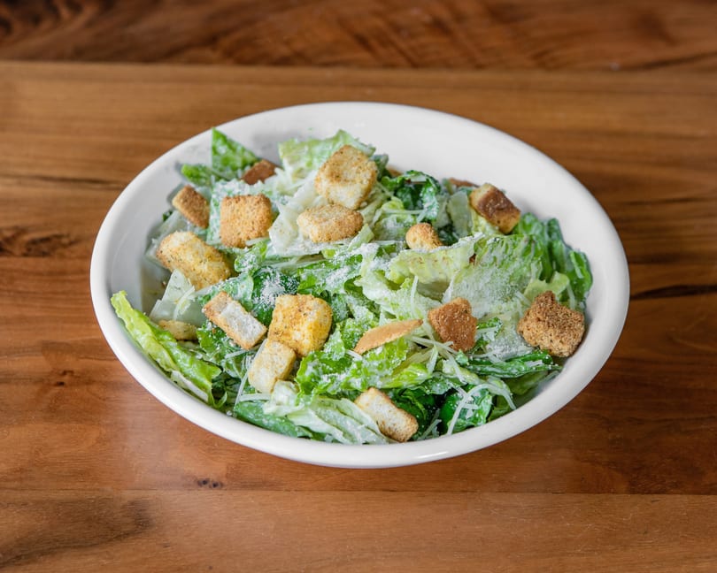 A white bowl filled with a Caesar salad, topped with croutons and grated cheese, on a wooden table.
