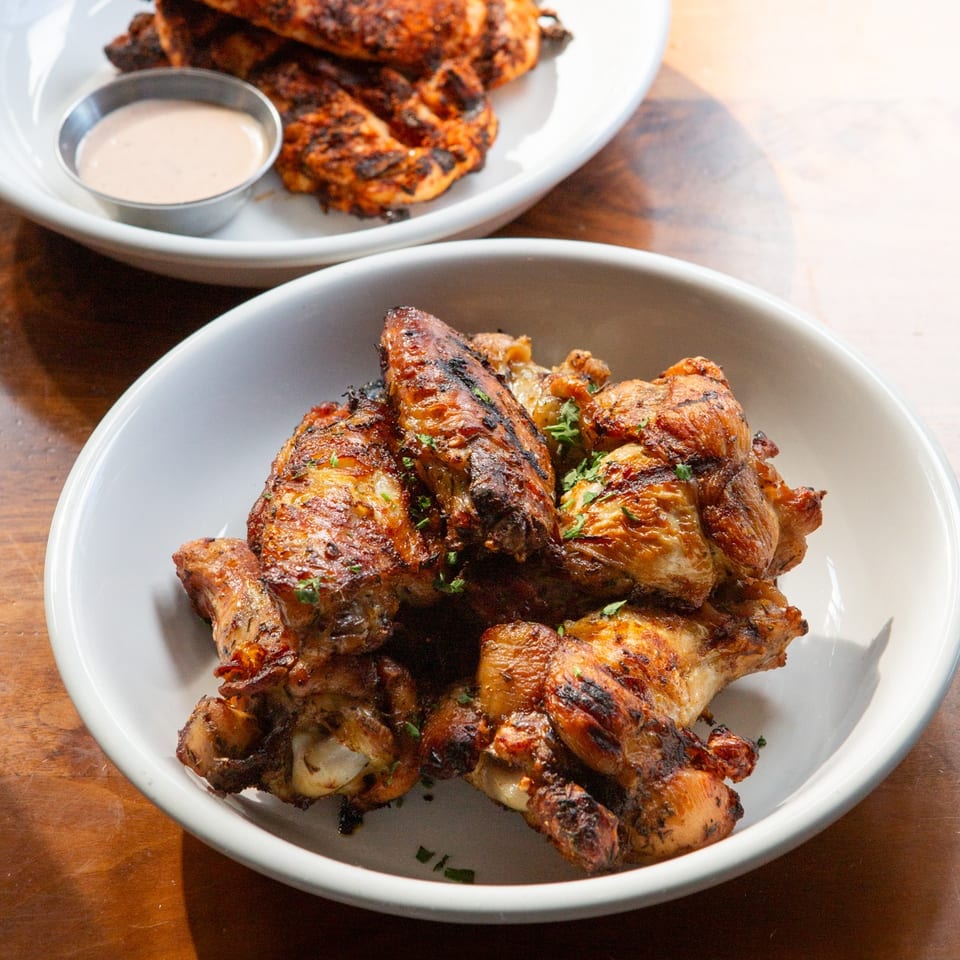 Two white bowls filled with grilled chicken wings and tenders, with a side of dipping sauce.