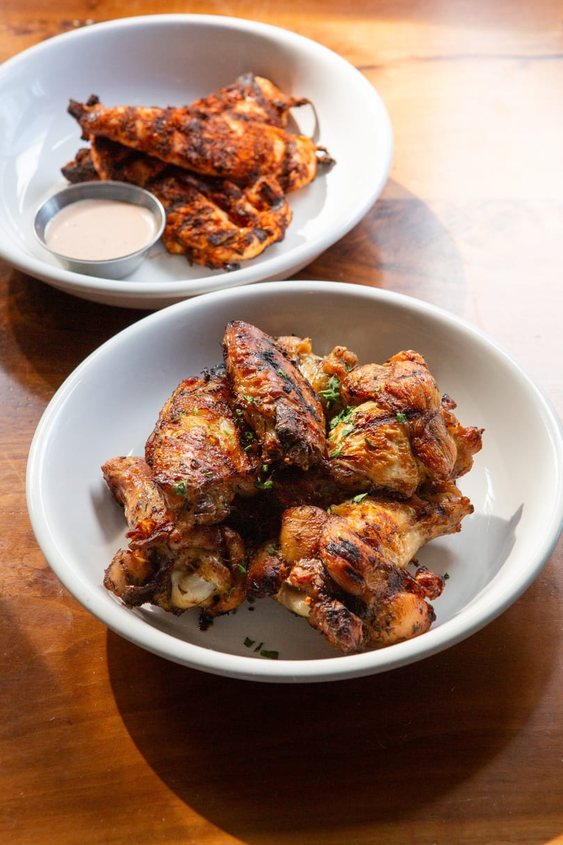 Two white bowls filled with grilled chicken wings and tenders, with a side of dipping sauce.